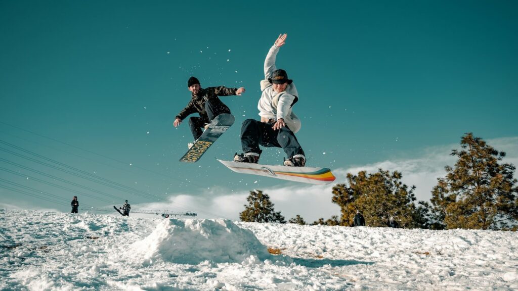 Twee snowboarders springen synchroon in de lucht boven een besneeuwde helling, voorbeeld van goede wintersport voorbereiding.